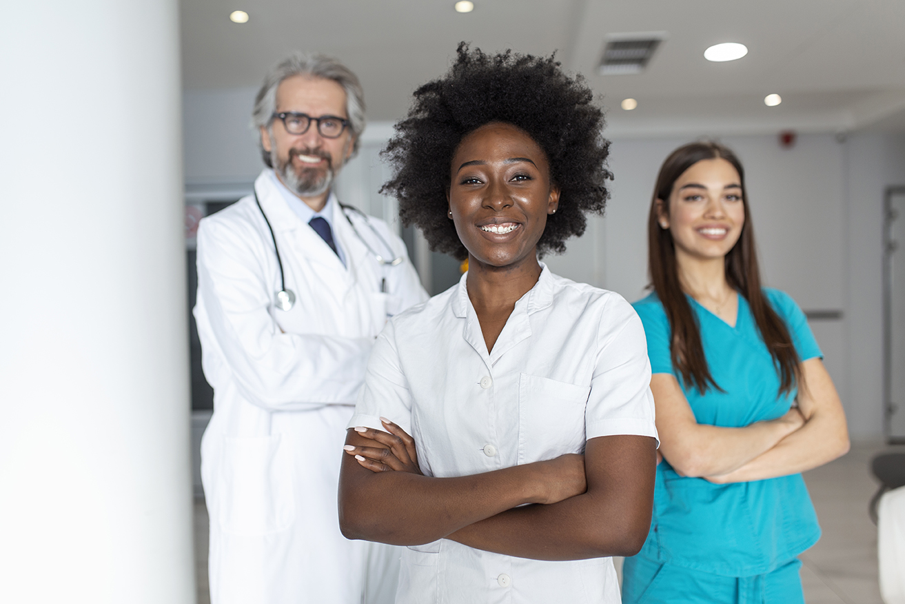 A multi-ethnic group of three doctors and nurses standing in a hospital corridor, wearing scrubs and coats. The team of healthcare workers are staring at the camera and smiling